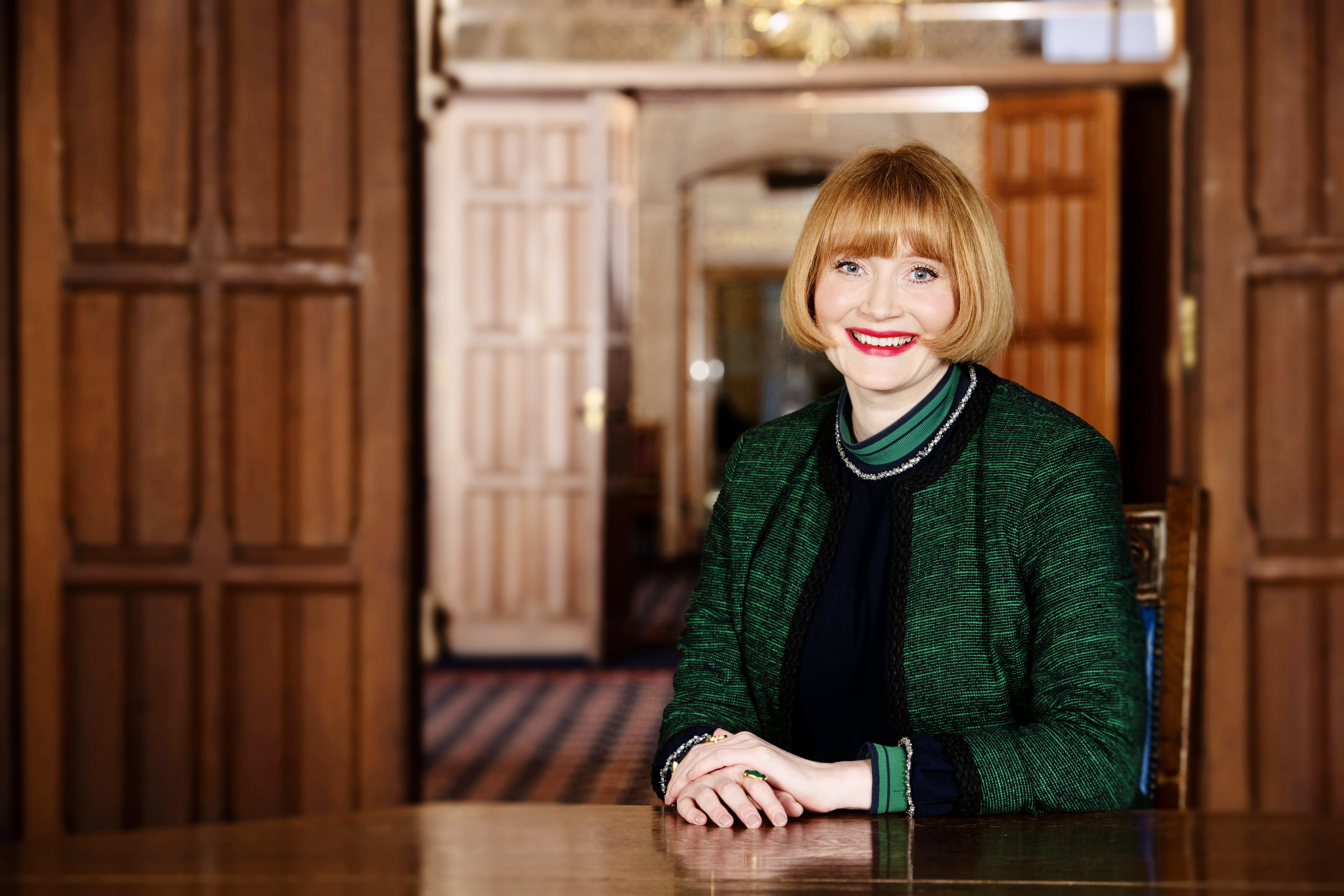 Women with a short blond bob smiles into the camera. She sits behind a large table in a dark wood panelled room.