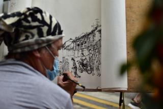 An aged street artist laying down a beautiful artwork on a paper canvas in public, painting down the street and sights around him.