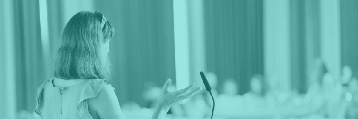 on left of image a woman with her back to us presents at a microphone to a lecture hall