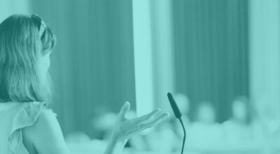 on left of image a woman with her back to us presents at a microphone to a lecture hall