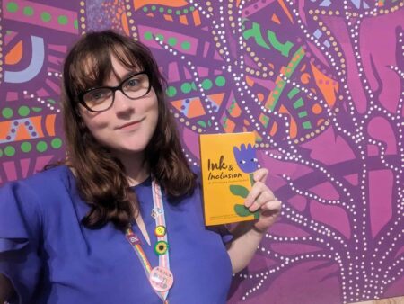 Brooke Szucs holding a book titled, "Ink and Inclusion" in front of a very colourful purple mural. 