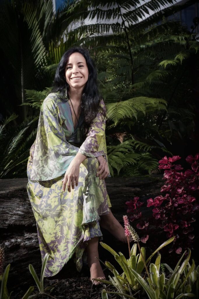 Photo of Gabriela Baron in leafy setting sat down and smiling to the camera