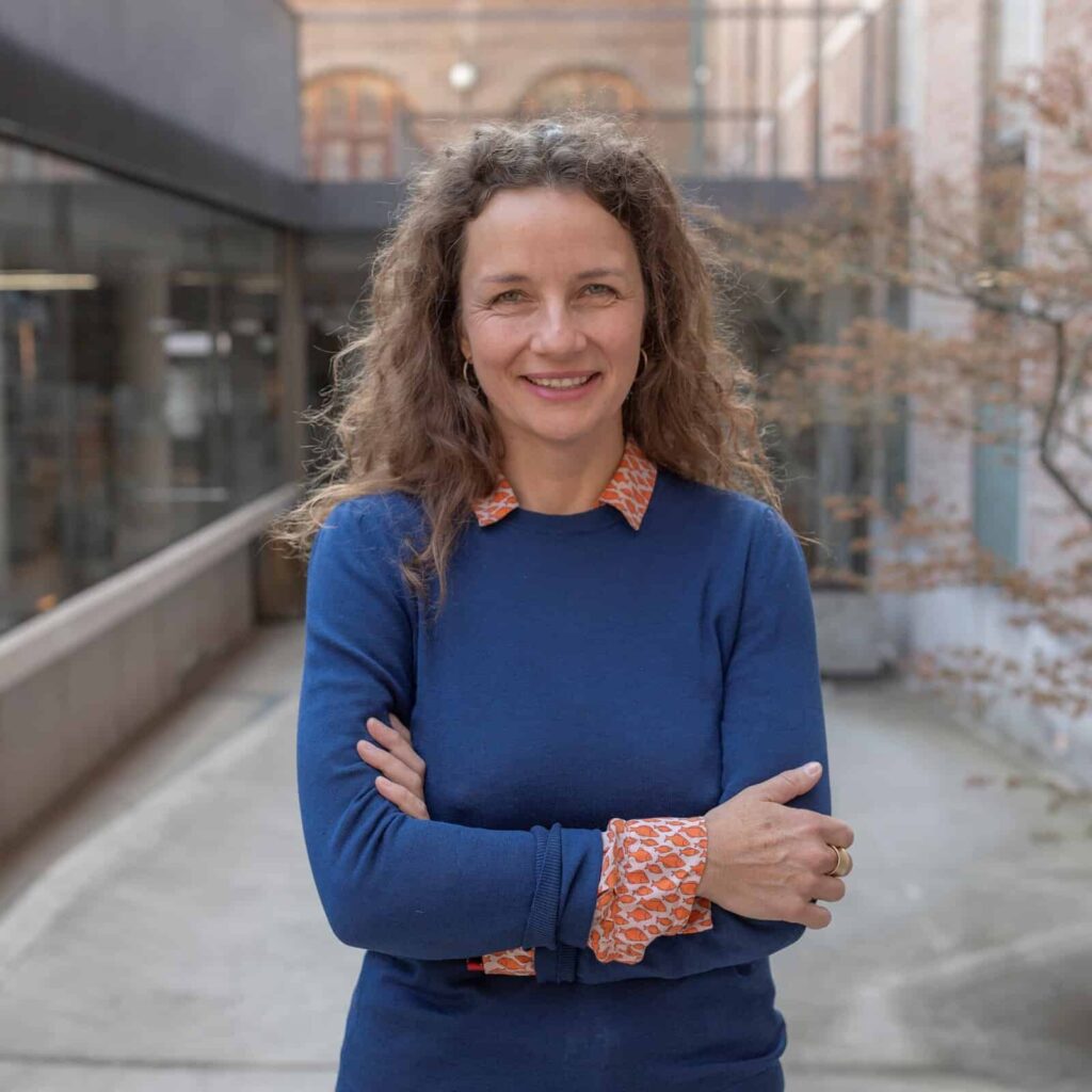 Photo of Paula on a campus setting smiling and facing the camera with arms crossed wearing a blue jumper and red shirt