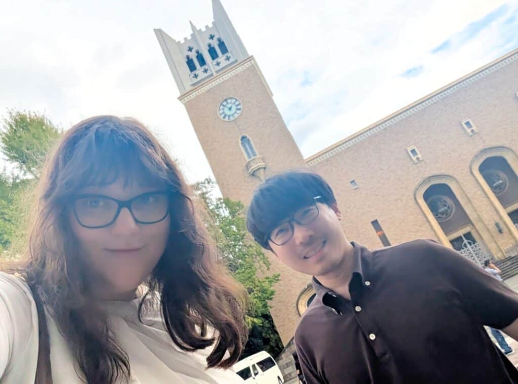 Image of Brooke Szucz and Shimpei Kumagai on campus at Waseda University standing in front of the Okuma Auditorium clock tower and smiling to the camera